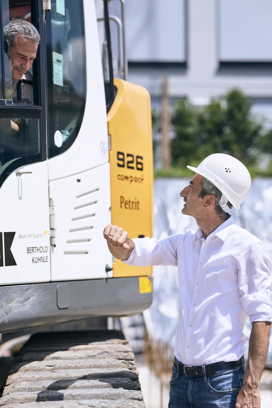 Ein Mann mit einem weißen Schutzhelm gestikuliert, während er mit einem Maschinenführer in einem Baufahrzeug auf einer Baustelle spricht.