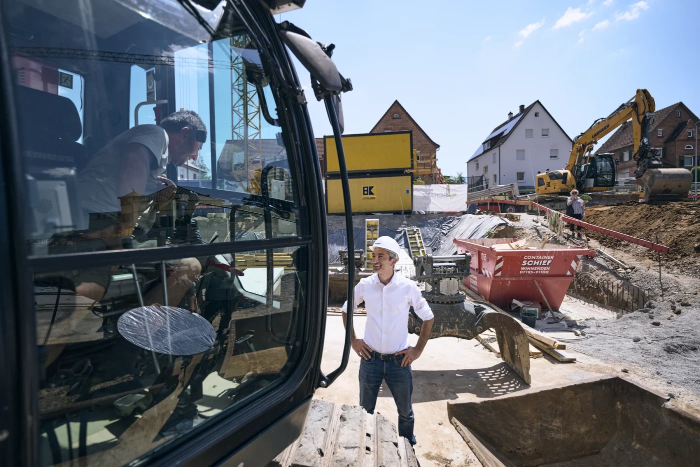 Eine Person in einem weißen Hemd und mit einem Schutzhelm steht auf einer Baustelle und beobachtet eine andere Person, die in einem großen Fahrzeug eine Maschine bedient. Im Hintergrund sind Erde, Baumaschinen und Häuser zu sehen.
