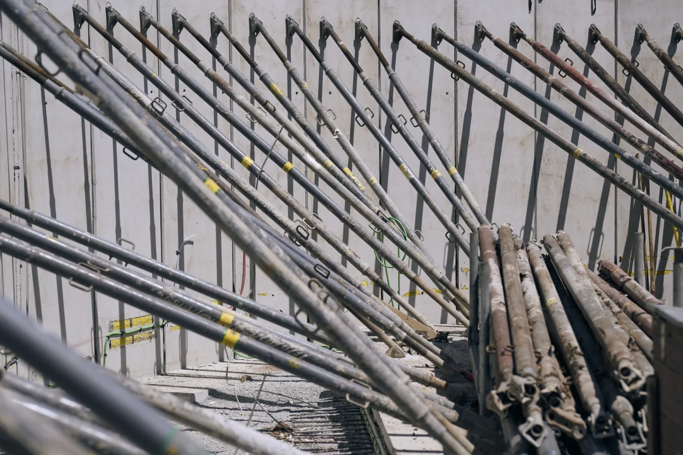 Eine Gruppe von Metallstangen und -stäben ist diagonal angeordnet und neben einer Wand auf einer Baustelle aufgestapelt, wodurch vor einem Betonhintergrund ein Muster aus sich kreuzenden Linien entsteht.