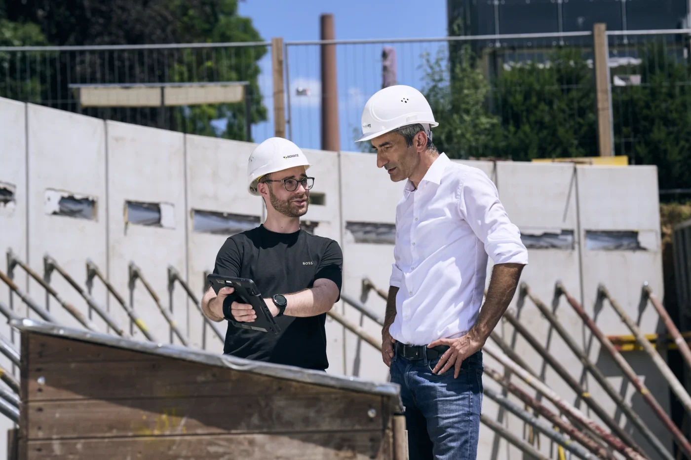 Zwei Männer mit weißen Schutzhelmen und in Geschäftskleidung stehen auf einer Baustelle. Der eine hält ein Tablet in der Hand und gestikuliert, während der andere zuhört; im Hintergrund sind Betonwände und Metallstangen zu sehen.